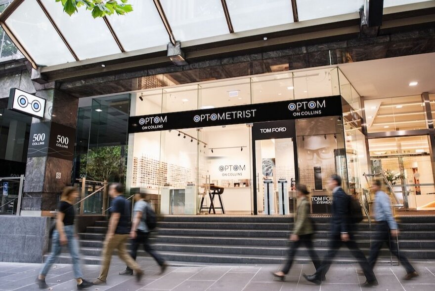 Pedestrians walking past a glass-fronted optometrist's shop with glasses on display inside.