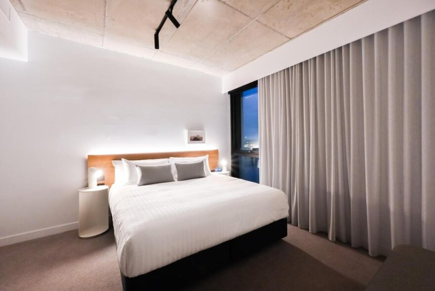 A bedroom at the Nesuto Docklands Apartment Hotel, with a king size bed, bedside tables and grey curtains.