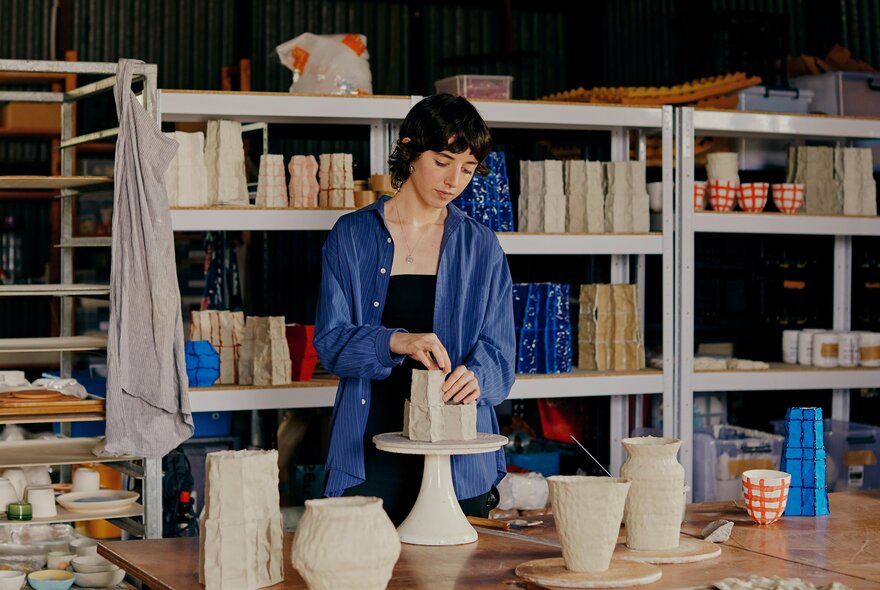 Ceramicist working on a pottery display with shelves of ceramics in the background.