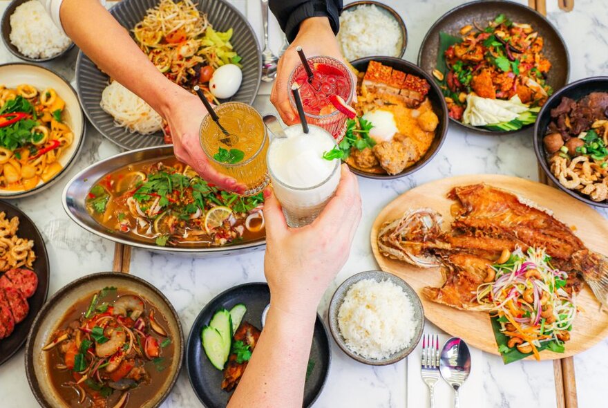 Hands clinking iced drinks with straws over a dining table with dishes of Thai food, stews, stir-fries and bowls of rice.