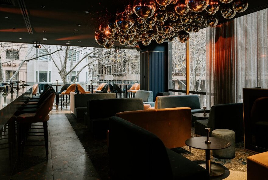 Interior of the Valiant, with soft velvet banquette seats and armchairs, a ceiling of golden glitter discos balls and the central bar area to the left of the image; large windows looking out onto the balcony and street.