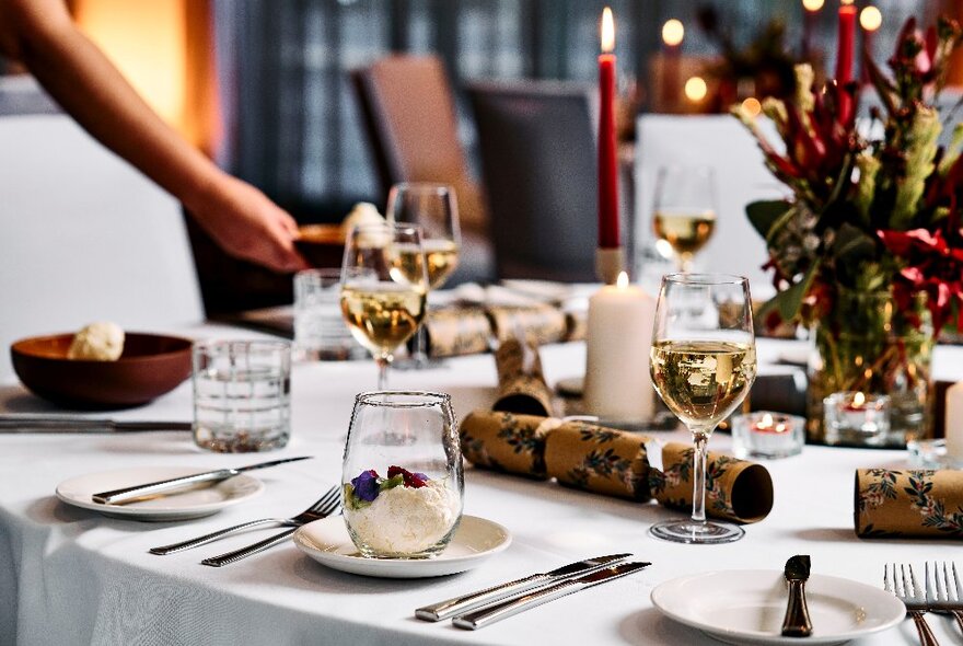 A waiter setting down a plate of food on a table set with white linen, silver cutlery, glasses of wine, festive decorations and candles.