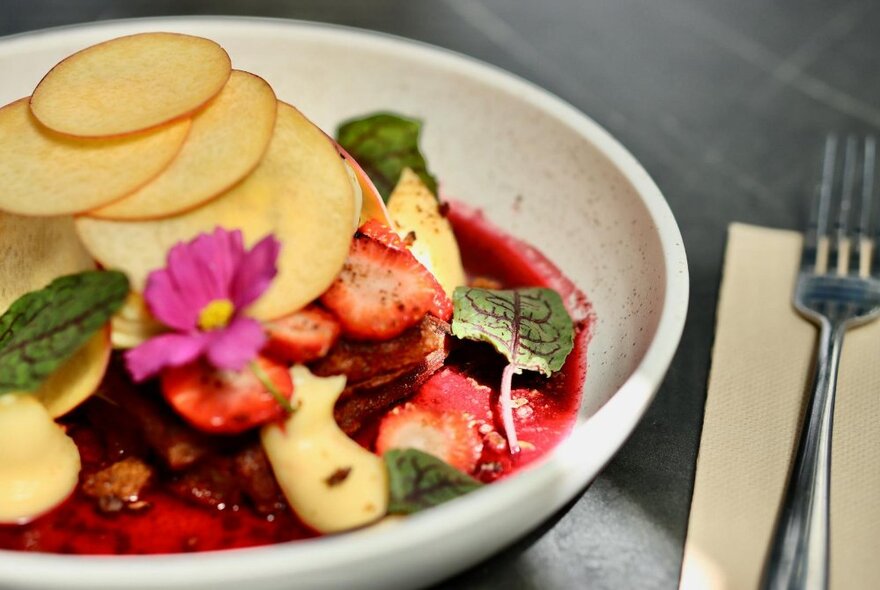 A dish of food including sliced apple and strawberries with a pink flower.