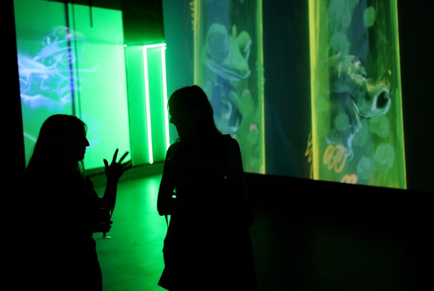 Two people in silhouette in a darkened exhibition space with images from the TV show Teenage Mutant Ninja Turtles projected onto the walls around them.