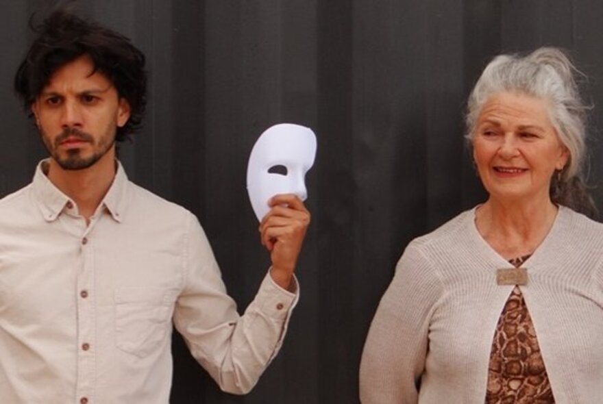 A man dressed in a beige shirt holding a white half-face mask standing next to an older woman in a beige cardigan.