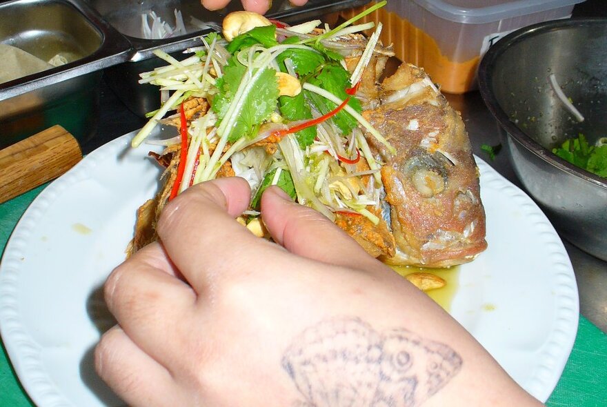 A hand with a butterfly tattoo, reaching into a dish of thai fried fish and salad. 
