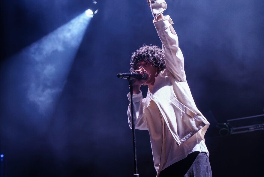 Singer performing on stage wearing a puffy jacket, holding onto a microphone stand with his arm raised under stage lights.