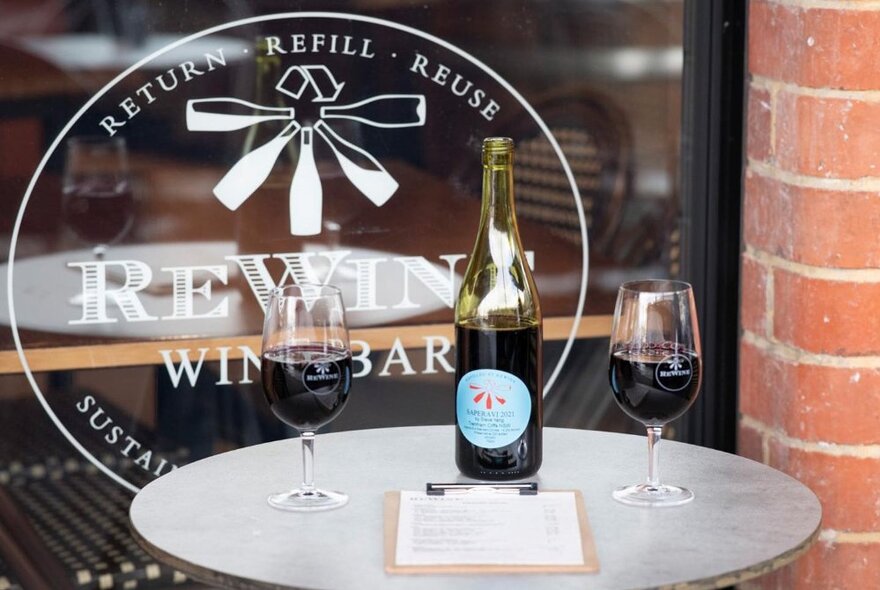 A round table outside the ReWine Wine Bar, with two full glasses and a half empty bottle of red wine, plus a clipboard with menu.