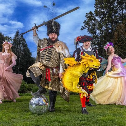 Four people in elaborate Renaissance and fantasy costumes, including a warrior with a sword on a disco ball, a man holding a yellow dragon, and two women in flowing gowns, pose on a lawn against a background of trees.
