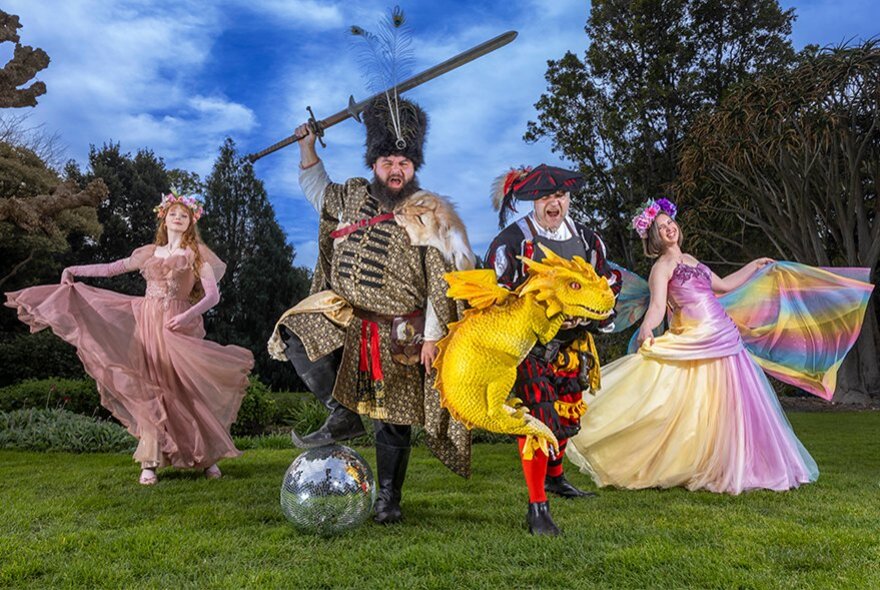 Four people in elaborate Renaissance and fantasy costumes, including a warrior with a sword on a disco ball, a man holding a yellow dragon, and two women in flowing gowns, pose on a lawn against a background of trees.