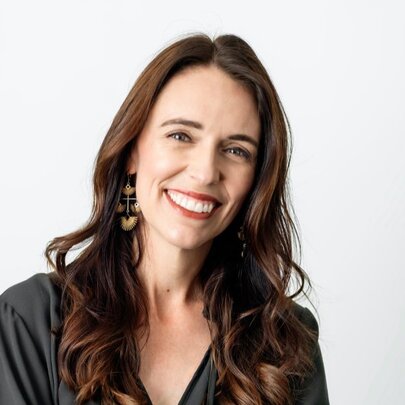 Jacinda Ardern smiling at camera, with her long brown hair framing her face, posed in front of a white background.