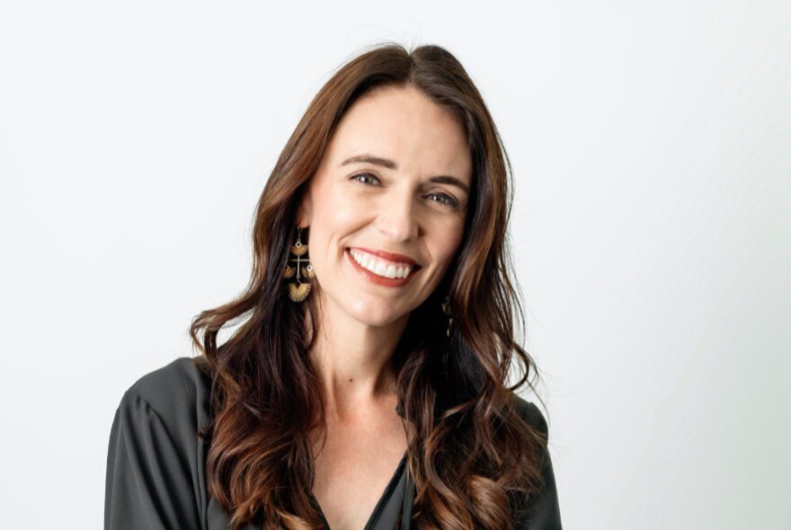 Jacinda Ardern smiling at camera, with her long brown hair framing her face, posed in front of a white background.