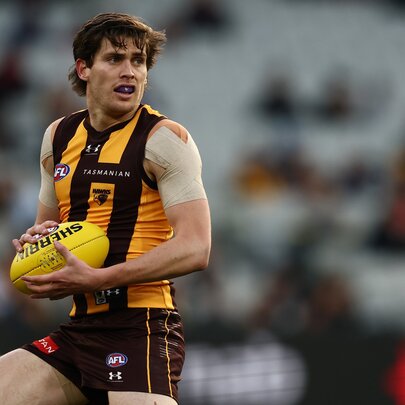 A Hawthorn AFL player running on the field while holding a football, blurred fans in the background.
