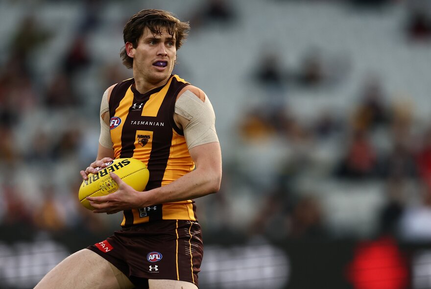 A Hawthorn AFL player running on the field while holding a football, blurred fans in the background.
