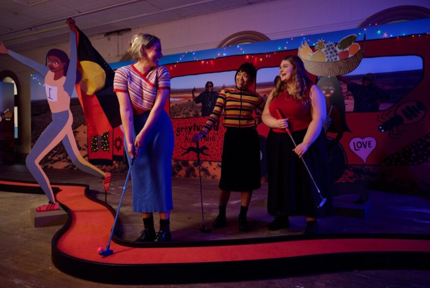 Three women wearing colourful outfits playing mini golf in a colourful indoor environment, with a red putting green.