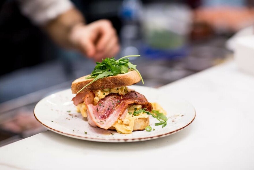 Bacon and egg sandwich on speckled white plate with hand out of focus in background, adding herbs.