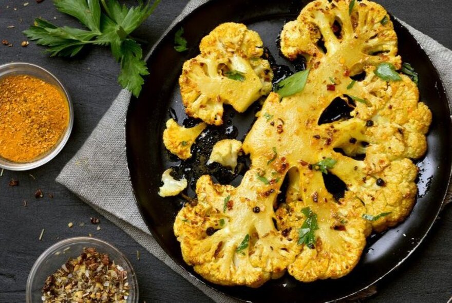 Grilled thick slices of cauliflower on a dark plate, with a small bowl of ground spices nearby.