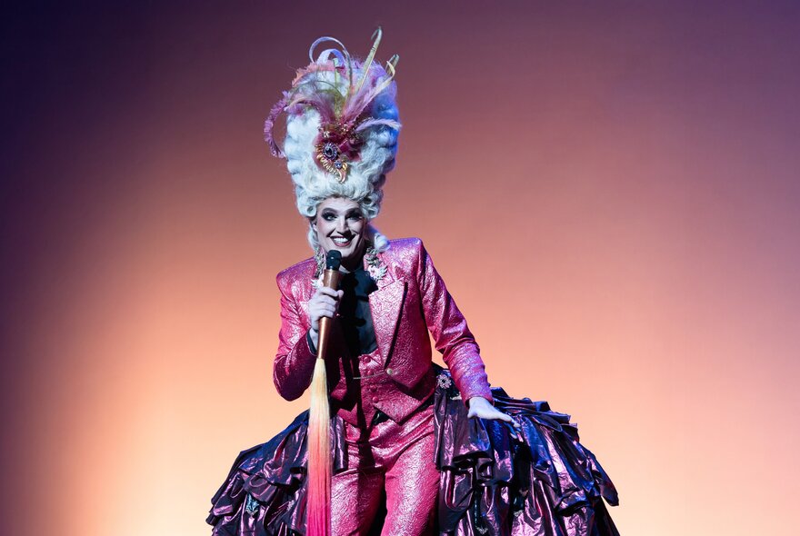 A person in a pink suit with a large headdress and heavy make-up, smiling and holding a microphone. 