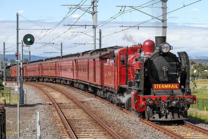 Red steam train chuffing along a track with wiring overhead.