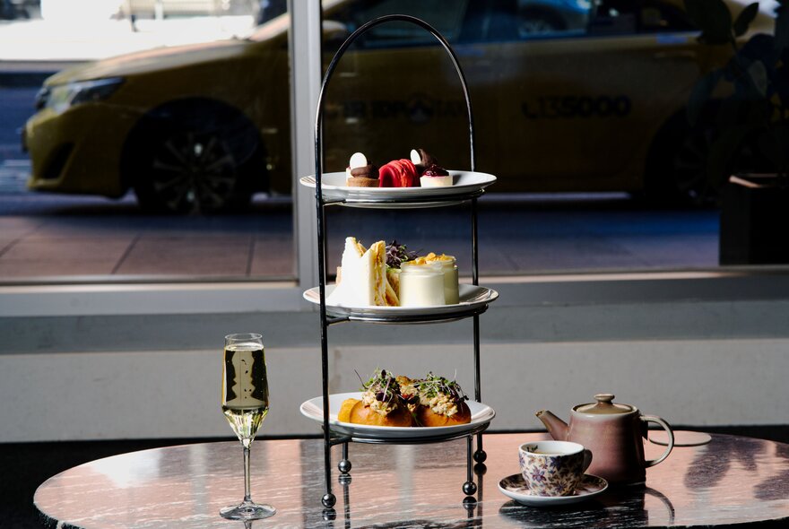 A table by a window with a car parked outside, set for High Tea with a three-tiered server. 