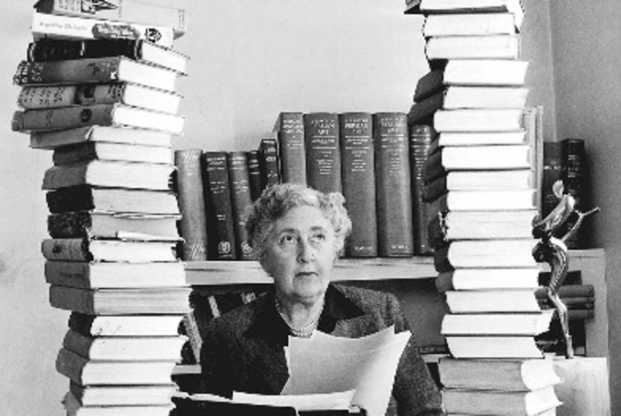 Agatha Christie seated at a table surrounded by towering piles of her many novels.