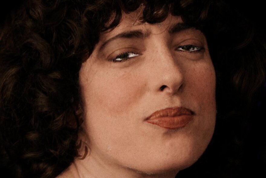 Close up of the face of comedian and actor Hannah Camilleri, she has slightly pursed lips and brown curly hair framing her face and the image appears to be hand coloured.