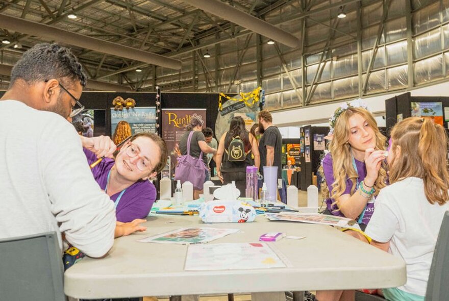 Two face painters in purple t-shirts painting the faces of two poeple at a trestle table in an exhibition setting.