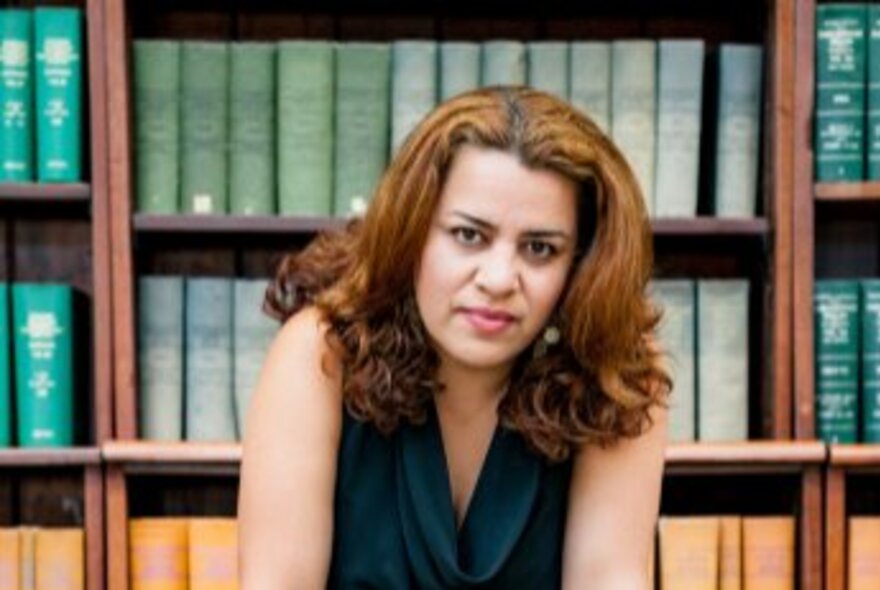 Portrait of Shokoofeh Azar, a woman with reddish-brown, wavy hair, wearing a dark top, in front of a bookshelf with numerous green and yellow-spined books.