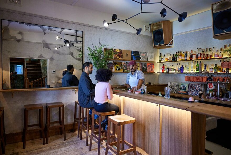 A couple sitting at the counter talking to a bartender over the bar, a large mirror on the side wall and vinyl record covers on display behind the bar.