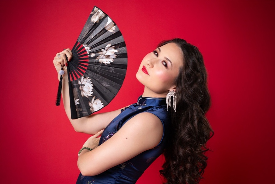 Comedian and cabaret artist Kelly Lim Harris holding an open fan and head tilted backwards against a red background.