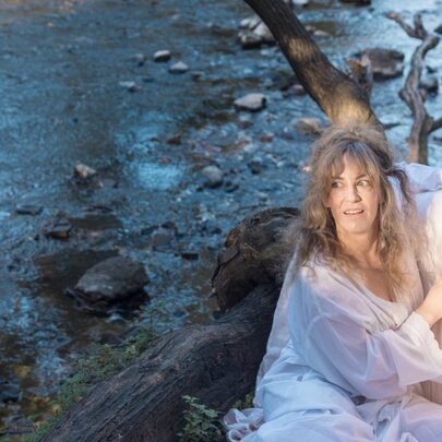 A woman with long, light-brown hair sits outdoors by a rocky stream.