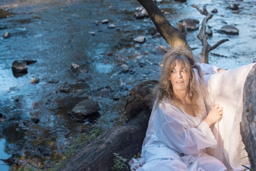 A woman with long, light-brown hair sits outdoors by a rocky stream.