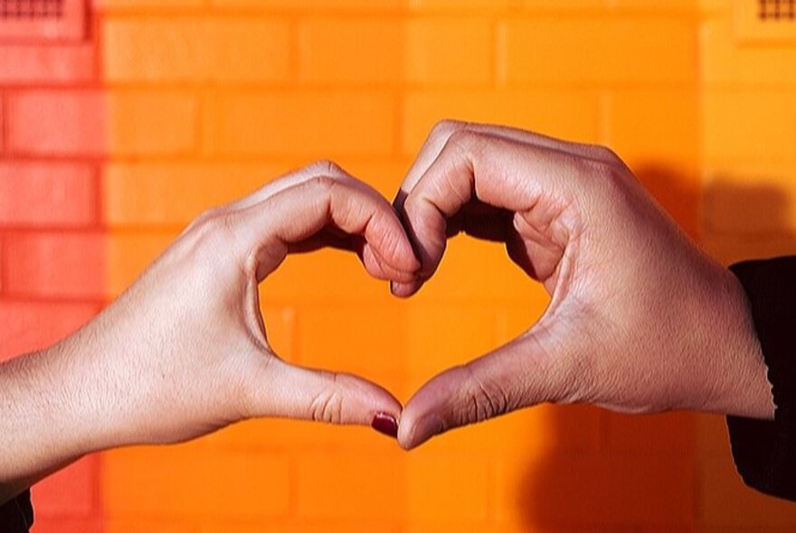 Two people's hands coming together to make the shape of a heart, in front of a rainbow-coloured brick wall.