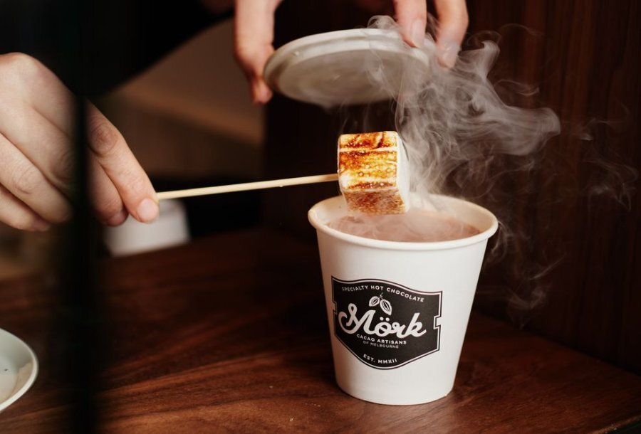 A woman putting a marshmallow into a steaming takeaway cup of hot chocolate.