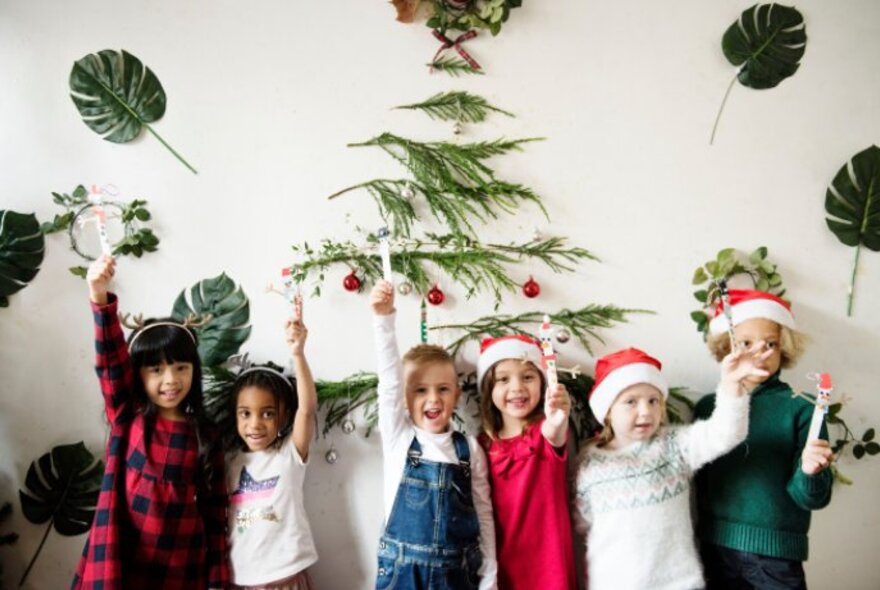 Children lined up against a white wall with Christmas decorations, wearing Santa hats and waving decorations.