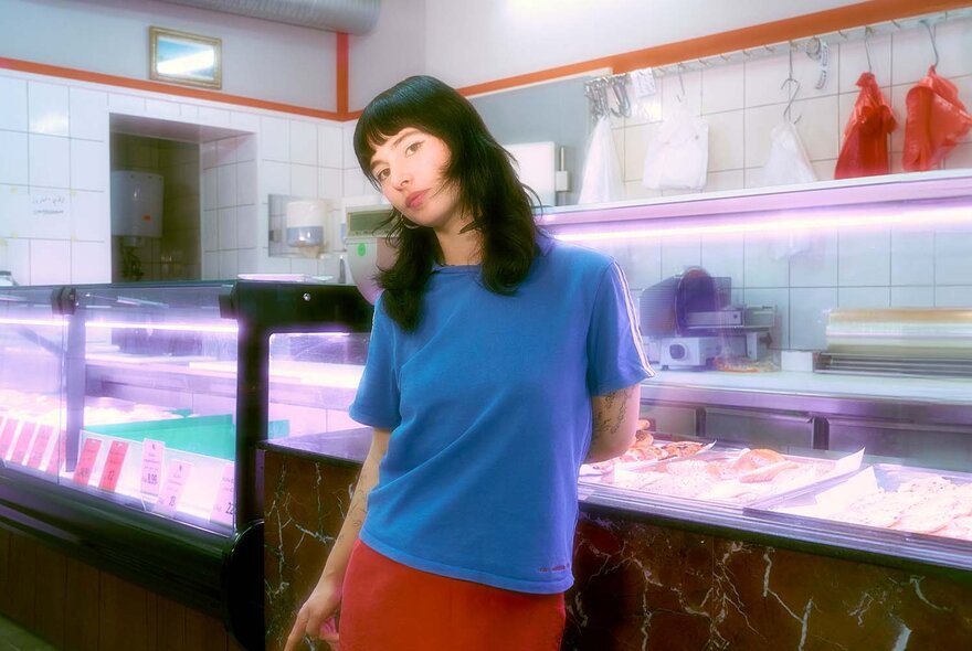 A soft-focus portrait of a woman in a blue t-shirt in front of a deli counter.