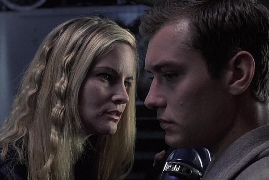 A scene from a film of a man looking blankly adn a woman with crimped blonde hair appearing to speak to him