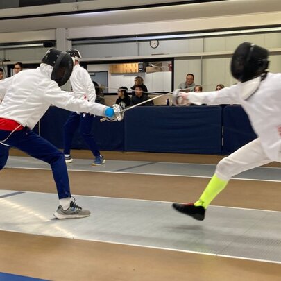 Two people dressed in white fencing outfits, fitted with head guards, fencing with swords on a fencing mat, indoors.