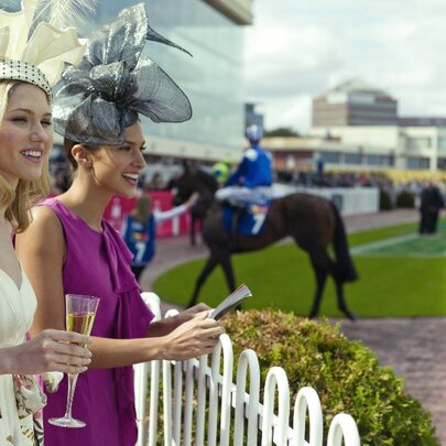 Two women (one holding a glass of champagne) dressed in frocks and hats at a race meeting.