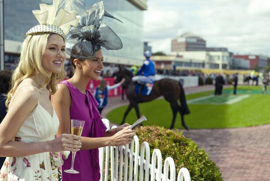 Two women (one holding a glass of champagne) dressed in frocks and hats at a race meeting.