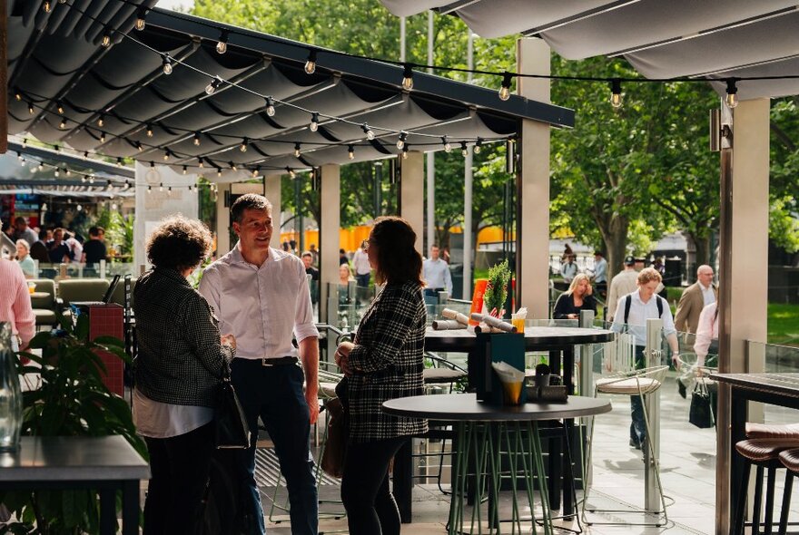 Three standing people enjoying a conversation outside a bar with others walking past in the background.