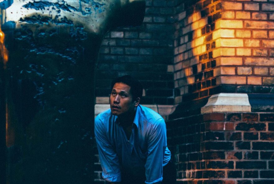 Musician Alex Zhang Hungtai, wearing a blue long-sleeved shirt and leaning forward slightly, his face looking upwards, with a brick building behind him.