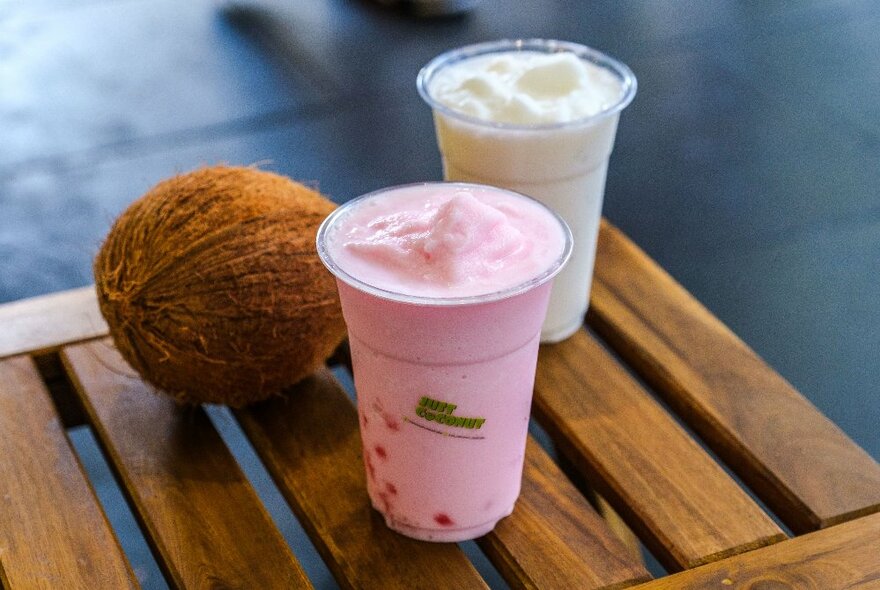 Two takeaway plastic cups filled with a coconut beverage, one white and one pink, with a whole coconut in the background. 