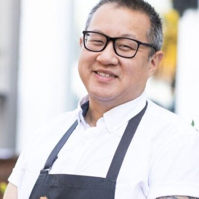 Chef Joe Hou, a smiling man, wearing a white chef shirt, a blue apron and glasses, standing outside.
