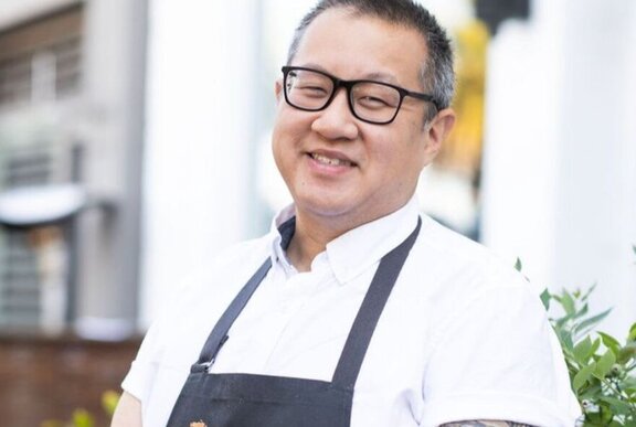 Chef Joe Hou, a smiling man, wearing a white chef shirt, a blue apron and glasses, standing outside.