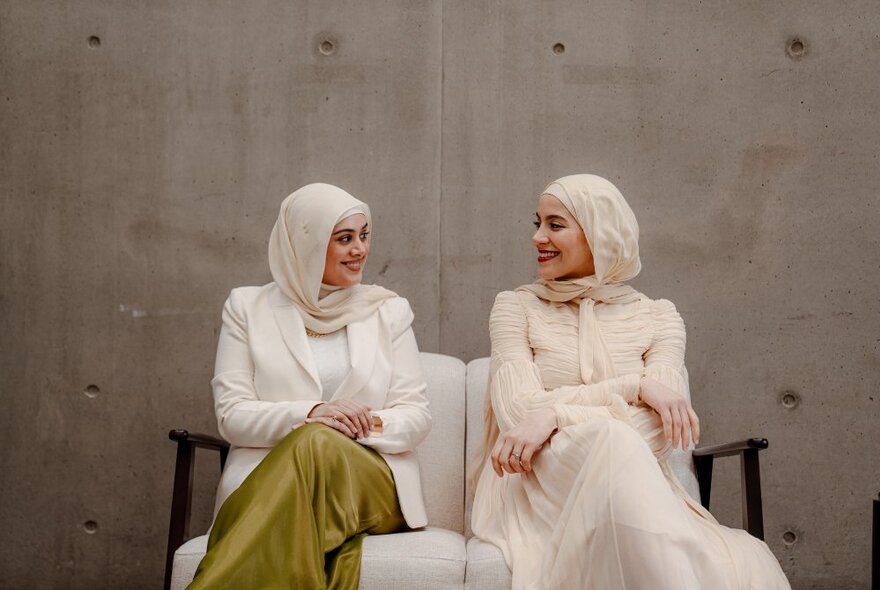 Two muslim women seated on a small couch in front of a concrete wall, smiling at each other.