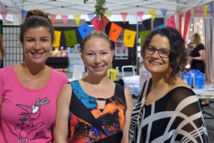 Three smiling women pose for a photo at an indoor market event with colorful pennant banners in the background.