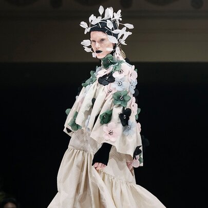 A fashion model wearing an avant-garde layered ensemble with floral embellishments and an elaborate headpiece from a runway collection.