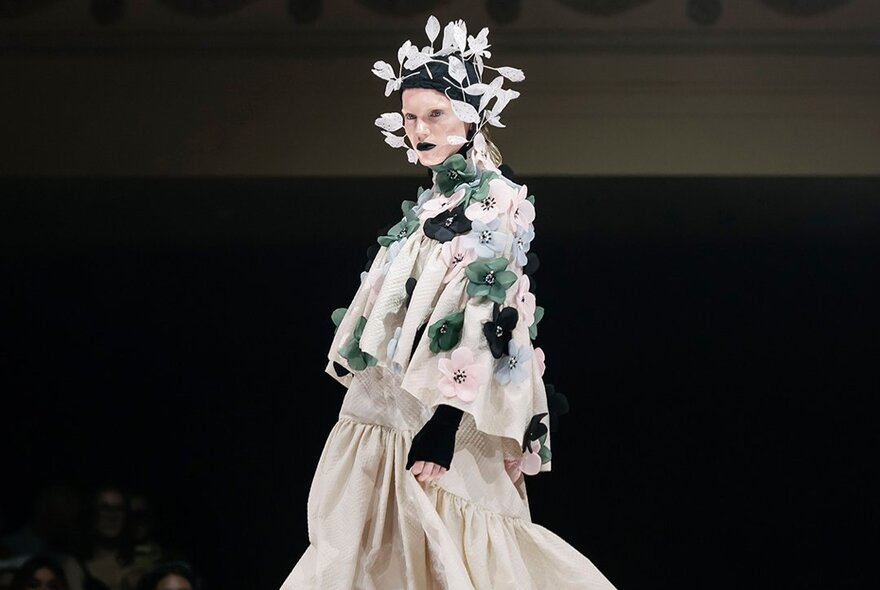 A fashion model wearing an avant-garde layered ensemble with floral embellishments and an elaborate headpiece from a runway collection.