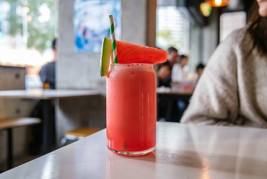 A watermelon juice on a table in a cafe with a slice of watermelon as garnish and a stripy straw. 